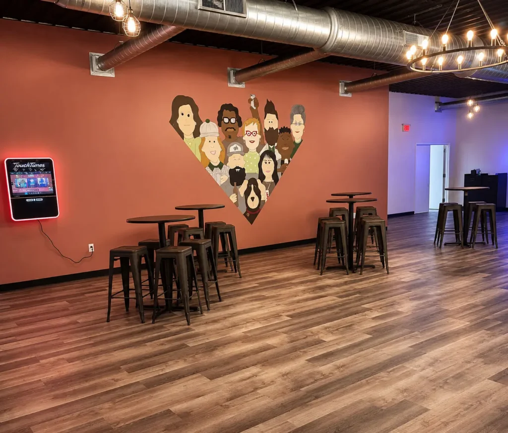 Open event space at Gathering Hub featuring high-top seating, decorative lighting, exposed ductwork, custom wall artwork, and flexible accommodations for celebrations, meetings, and community events.